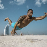 A focused male fitness athlete performing an exercise on the sunny white sand beach of Dubai, with the iconic Burj Al Arab hotel in the background.