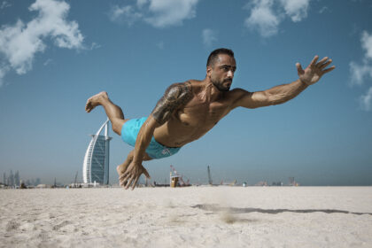A focused male fitness athlete performing an exercise on the sunny white sand beach of Dubai, with the iconic Burj Al Arab hotel in the background.