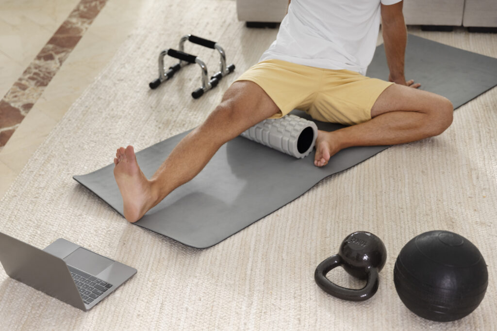 A man performing self-myofascial release by foam rolling his hamstring muscle on a yoga mat at home.