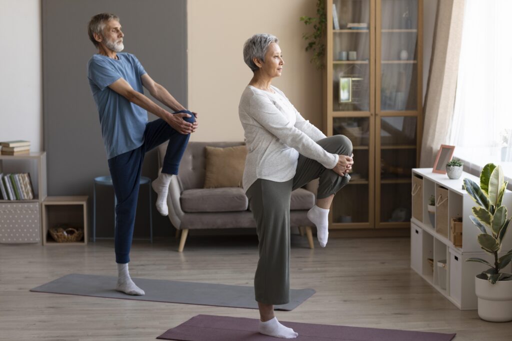 Lifespan fitness: A senior couple at home performing a balance and flexibility exercise, each standing on one leg while stretching their hamstring.