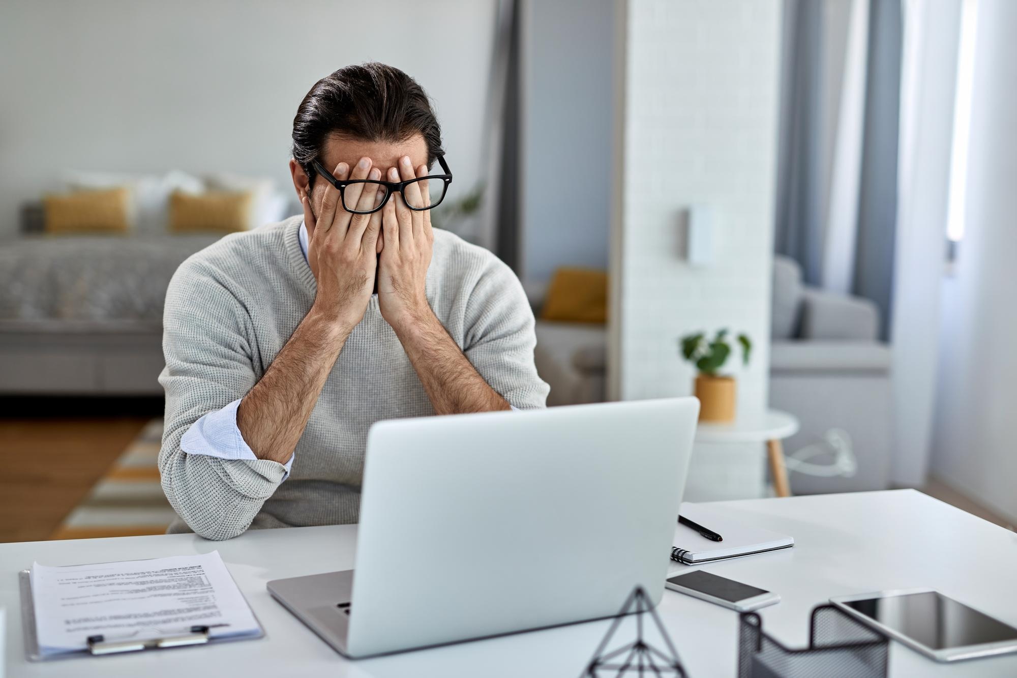 A stressed man at his desk in a modern office, covering his eyes in exhaustion while his laptop screen glows with unfinished work.