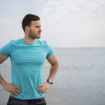 A confident, healthy man standing on a shoreline, looking out to sea with his hands on his waist, embodying vitality and a balanced lifestyle.