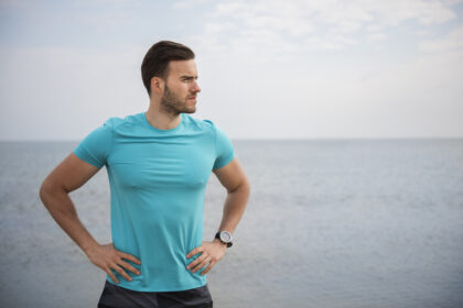 A confident, healthy man standing on a shoreline, looking out to sea with his hands on his waist, embodying vitality and a balanced lifestyle.