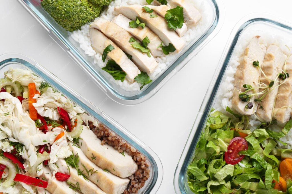 A top-down view of a weekly healthy meal prep container with grilled chicken breast, rice, and a fresh green salad.