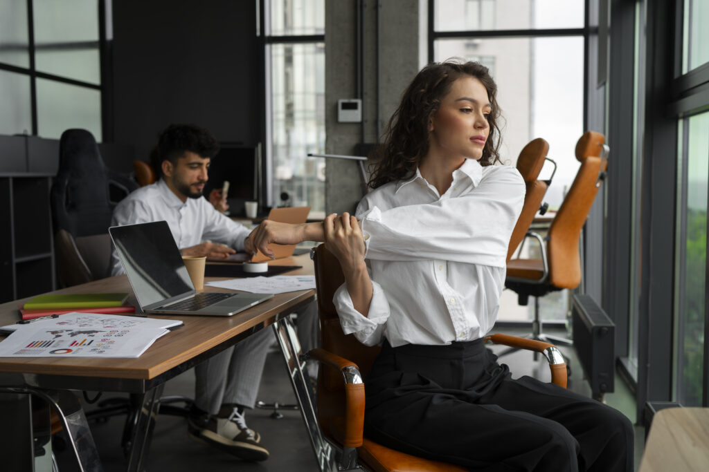 A young woman in an office taking an active break, performing a simple seated stretch and breathing exercise at her desk.