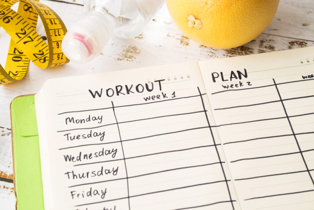 A top-down shot of a fitness planner open to "Workout week 1" and "Plan week 2," with days of the week listed. A yellow measuring tape, a clear water bottle with a pink cap, and a grapefruit are visible in the background, all resting on a distressed white wooden surface.